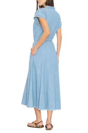 A woman stands with her back turned, wearing the Xirena Dixie Chambray Dress—a light blue, short-sleeve, button-front cotton midi dress—paired with leopard print sandals.