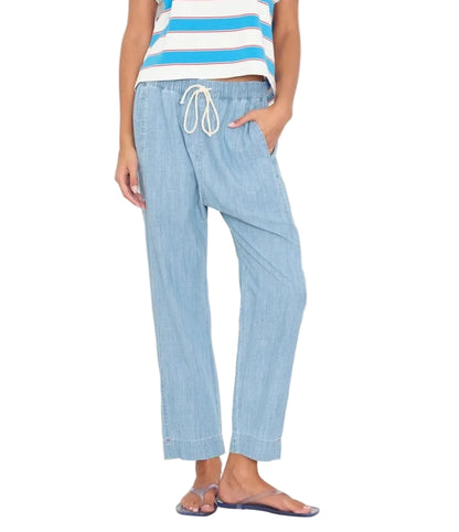 Wearing the Xirena Draper Chambray Pant in light blue with a drawstring waist, a striped sleeveless top, and sandals, the person stands with one hand in their pocket.
