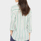 A woman with long brown hair stands facing away, wearing a modern silhouette Xirena Bowie Stripe Shirt—a long-sleeve, multicolored striped viscose-linen shirt—paired with dark pants.
