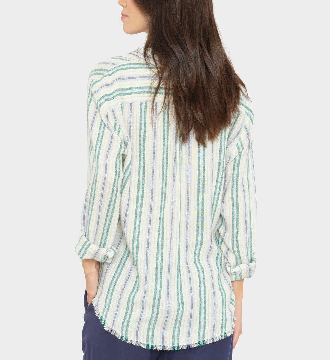 A woman with long brown hair stands facing away, wearing a modern silhouette Xirena Bowie Stripe Shirt—a long-sleeve, multicolored striped viscose-linen shirt—paired with dark pants.
