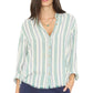 A woman is wearing the Xirena Bowie Stripe Shirt—a modern button-up shirt featuring long sleeves rolled up and bold vertical green, blue, and white stripes in a viscose-linen blend. The shirt is partially tucked into dark blue pants.