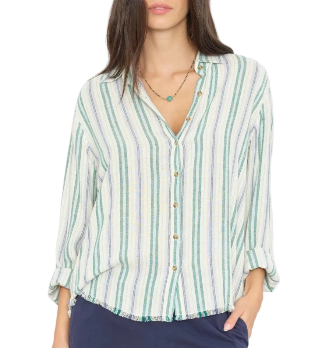A woman is wearing the Xirena Bowie Stripe Shirt—a modern button-up shirt featuring long sleeves rolled up and bold vertical green, blue, and white stripes in a viscose-linen blend. The shirt is partially tucked into dark blue pants.