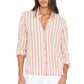 A woman wears the Xirena Bowie Stripe Shirt—a modern, long-sleeve button-up featuring vertical pink and white stripes in a soft viscose-linen blend. Designed with a collar and rolled cuffs, the shirt is styled untucked with crisp white bottoms.