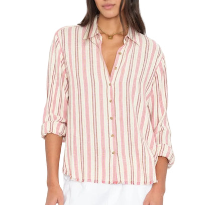 A woman wears the Xirena Bowie Stripe Shirt—a modern, long-sleeve button-up featuring vertical pink and white stripes in a soft viscose-linen blend. Designed with a collar and rolled cuffs, the shirt is styled untucked with crisp white bottoms.