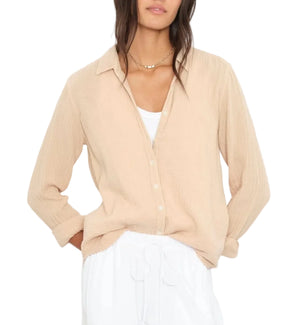 A woman wears the Xirena Scout Cotton Gauze Shirt in beige over a white top and white drawstring pants, hands in her pockets.