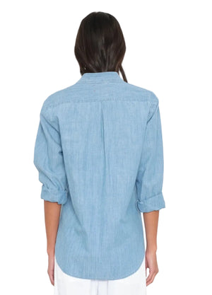 A person stands with their back to the camera wearing the Xirena Beau Chambray Shirt in light blue, styled with rolled-up sleeves and paired with white pants.