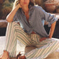 Woman sits outdoors on stone, wearing the Xirena Beau Chambray Shirt with striped wide-leg pants and brown sandals; plants and pottery appear in the background.