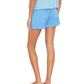 A person faces away, wearing Xirena’s Declan Short—a lightweight cotton poplin pull-on with an elasticized drawstring waist—paired with a blue checkered top and blue sandals against a white background.