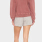A woman faces away from the camera, wearing Xirena’s Zane Short in herringbone striped cotton with a pink long-sleeve top and black sandals, posed against a plain light background.