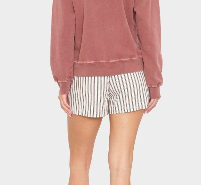 A woman faces away from the camera, wearing Xirena’s Zane Short in herringbone striped cotton with a pink long-sleeve top and black sandals, posed against a plain light background.