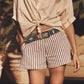 A person stands outdoors among plants wearing Xirena's Zane Short—tailored mid-rise herringbone striped cotton shorts with a blue patterned belt—paired with a beige button-up shirt tied at the waist.