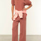 A person wears Xirena's Matteo Sweatpant in mauve, a vintage-washed French terry wide-leg cropped style with a matching top, light pink sweatshirt tied at the waist, and brown slippers, standing against a plain background.
