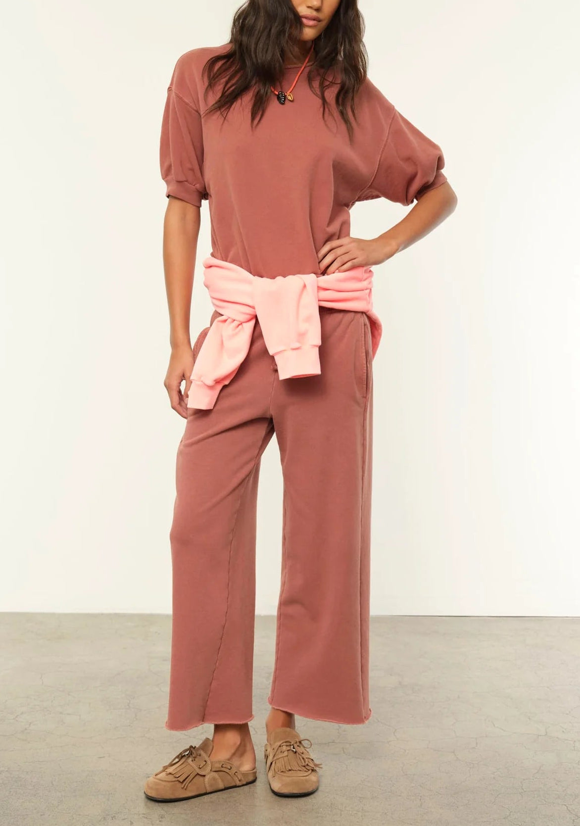 A person wears Xirena's Matteo Sweatpant in mauve, a vintage-washed French terry wide-leg cropped style with a matching top, light pink sweatshirt tied at the waist, and brown slippers, standing against a plain background.