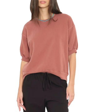 A woman wears the Xirena Jax Sweatshirt, a loose dusty rose short-sleeve top in vintage washed French terry, paired with black drawstring pants, standing against a white background.