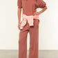Wearing the Xirena Jax Sweatshirt in a loose, rose vintage-wash style with matching wide-leg pants and tan sneakers, this person has a light pink sweatshirt tied at the waist and stands indoors.
