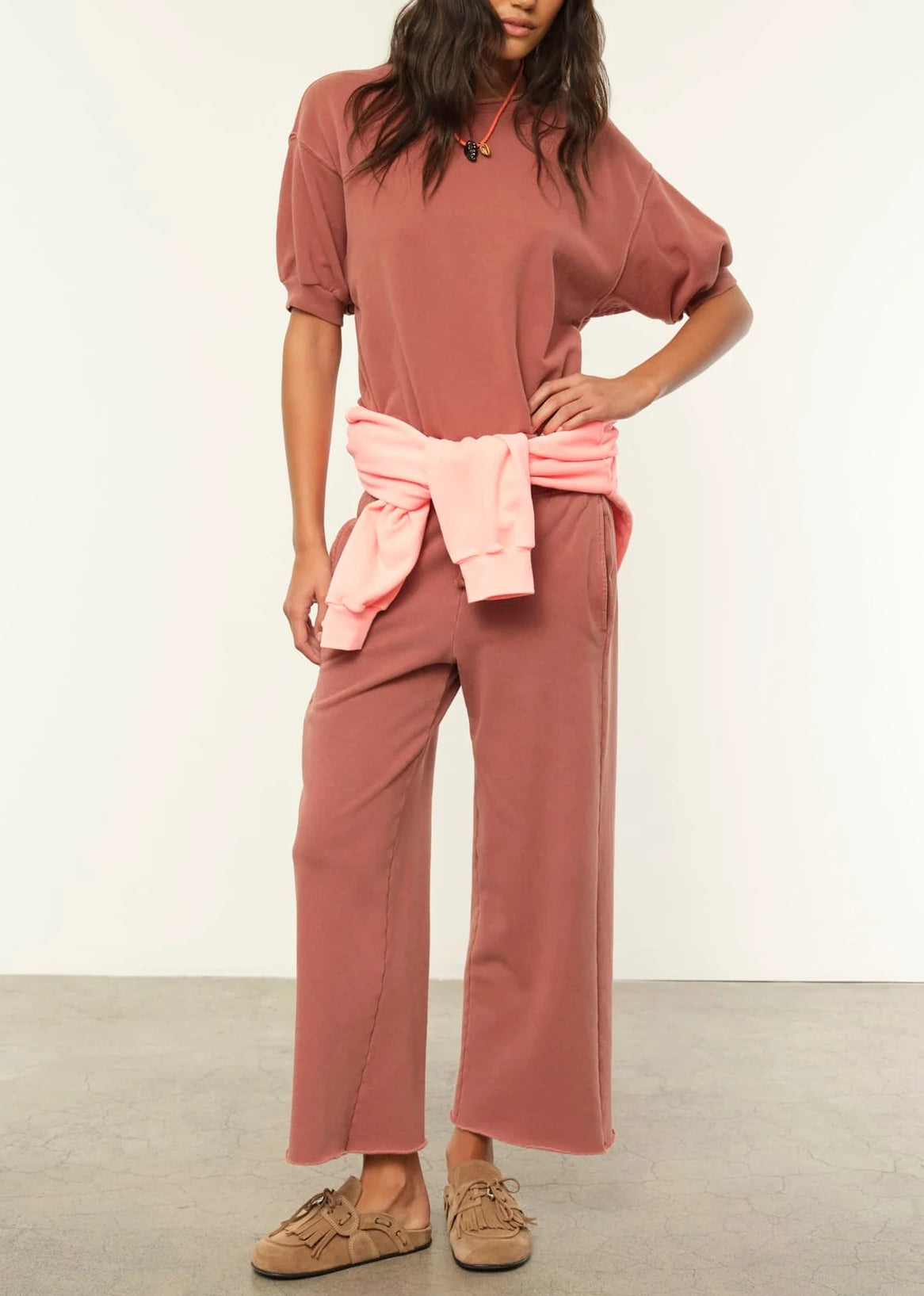 Wearing the Xirena Jax Sweatshirt in a loose, rose vintage-wash style with matching wide-leg pants and tan sneakers, this person has a light pink sweatshirt tied at the waist and stands indoors.