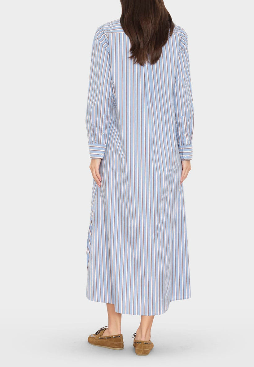 A woman stands with her back to the camera, wearing the Boden Dress by Xirena—a long-sleeved, blue and white vertical striped tailored shirtdress—paired with brown loafers.