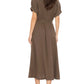 A woman is wearing the Xirena Cate Dress—a brown, short-sleeve cotton gauze dress featuring an adjustable tie belt and A-line skirt—paired with black flat shoes, standing and facing away.
