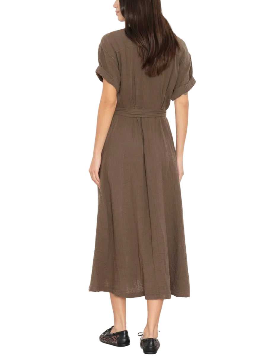 A woman is wearing the Xirena Cate Dress—a brown, short-sleeve cotton gauze dress featuring an adjustable tie belt and A-line skirt—paired with black flat shoes, standing and facing away.