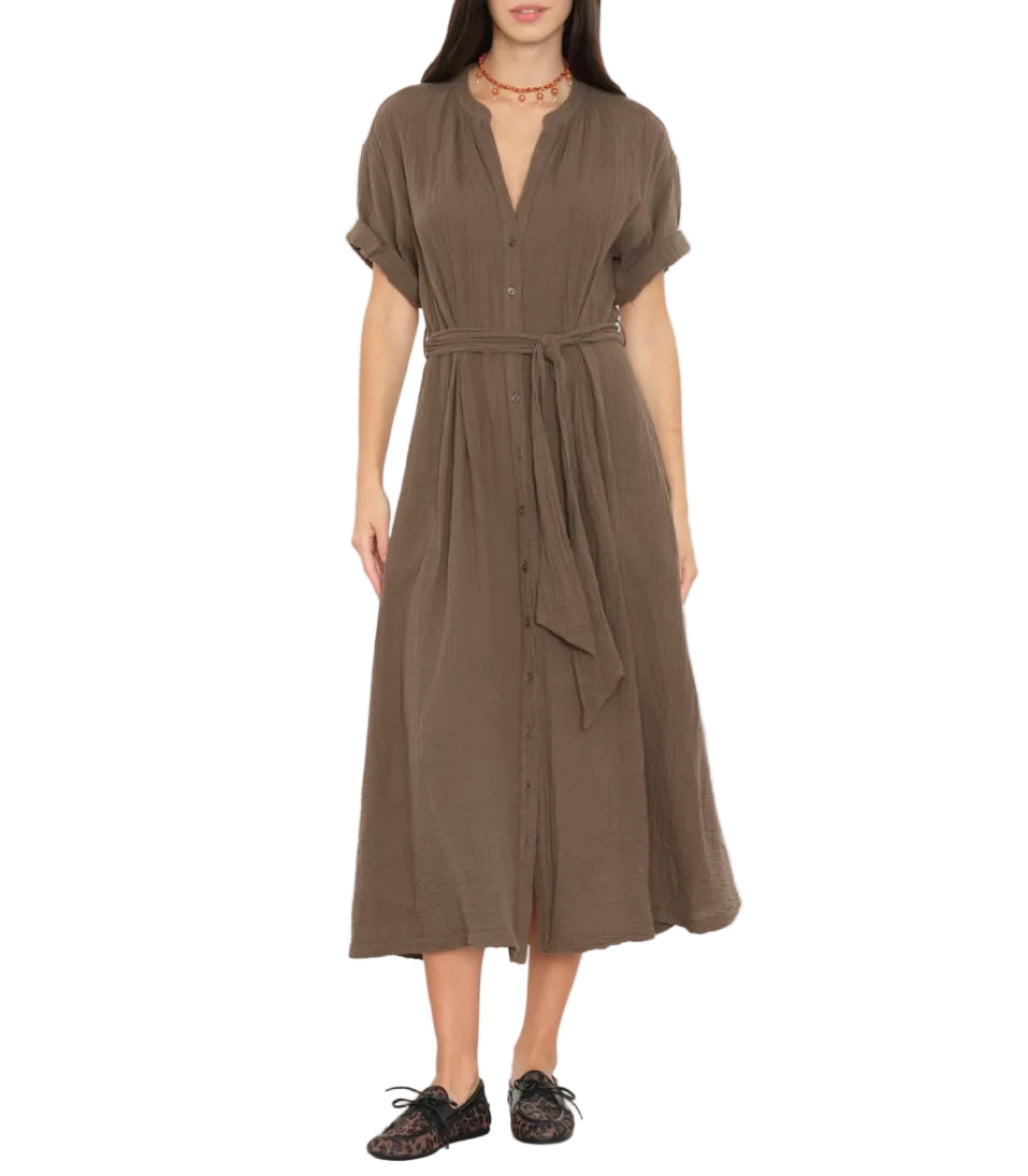 A woman stands against a white background wearing the Xirena Cate Dress—a brown, short-sleeve, button-down cotton gauze dress with an adjustable tie belt—paired with black shoes and a red beaded necklace.