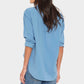 A woman, facing away, wears the Xirena Beau Shirt—a loose, menswear-inspired light blue cotton poplin shirt with rolled sleeves—paired with blue jeans against a plain light background.
