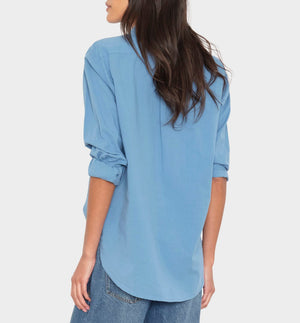 A woman, facing away, wears the Xirena Beau Shirt—a loose, menswear-inspired light blue cotton poplin shirt with rolled sleeves—paired with blue jeans against a plain light background.