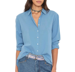A woman wears the Xirena Beau Shirt—a blue cotton poplin button-up styled with rolled-up sleeves—partially tucked into blue jeans. She completes her effortlessly menswear-inspired look with a beaded belt and a black beaded choker necklace.