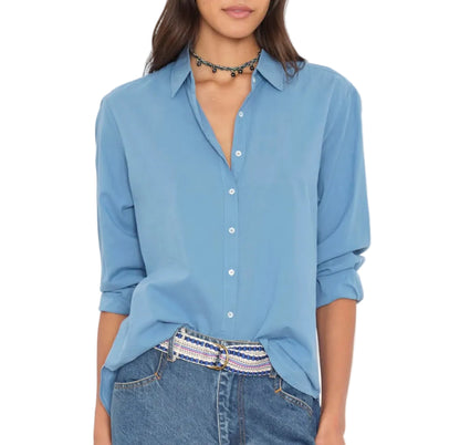 A woman wears the Xirena Beau Shirt—a blue cotton poplin button-up styled with rolled-up sleeves—partially tucked into blue jeans. She completes her effortlessly menswear-inspired look with a beaded belt and a black beaded choker necklace.