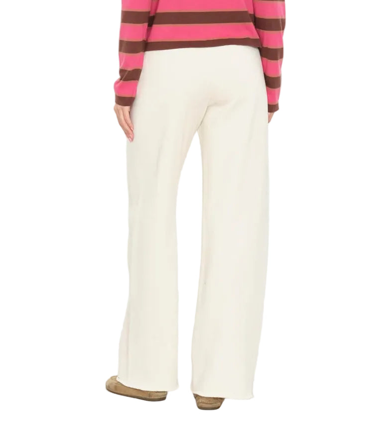 A person is shown from the back against a plain white background wearing the Xirena Renzo Sweatpant in white, featuring a wide-leg silhouette and an elasticized drawstring waist, paired with brown shoes and a pink and brown striped long-sleeve top.