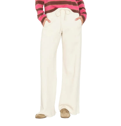 Wearing the Xirena Renzo Sweatpant—a cream-colored, wide-leg style with an elasticized drawstring waist—paired with tan shoes and a pink and brown striped long-sleeve shirt, hands in pockets, photographed from shoulders down.