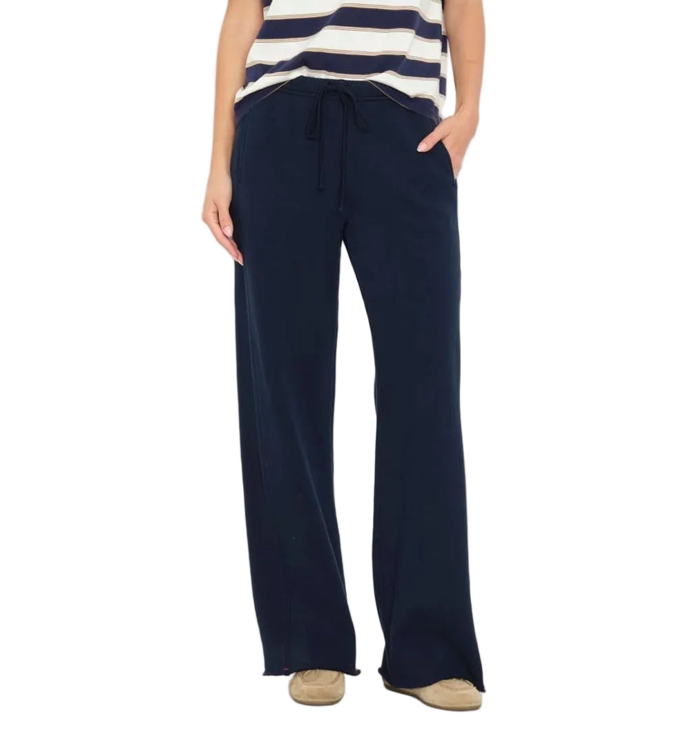 A person wearing the Xirena Renzo Sweatpant in navy with an elasticized drawstring waist, styled with a striped short-sleeve top and beige shoes, photographed from the shoulders down against a white background.