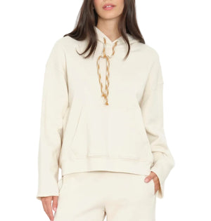 A woman wearing the Xirena Chase Sweatshirt, a relaxed hoodie made from ultra-soft fleece with a kangaroo pocket and beige drawstrings, paired with matching off-white pants.