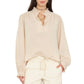 A woman wearing the Xirena Dorien Top—a light beige, lightweight cotton poplin long-sleeve blouse featuring blouson sleeves and ruffled collar and cuffs—paired with matching drawstring pants, stands with one hand in her pocket.