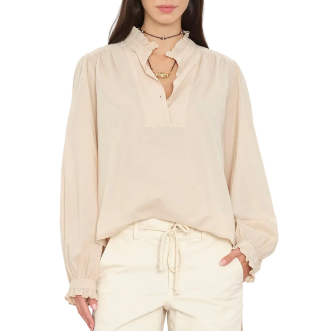 A woman wearing the Xirena Dorien Top—a light beige, lightweight cotton poplin long-sleeve blouse featuring blouson sleeves and ruffled collar and cuffs—paired with matching drawstring pants, stands with one hand in her pocket.