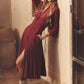 A woman stands against a stucco wall, one hand in her pocket and the other resting above her head, wearing the Xirena Louisa Dress—a long-sleeved maroon button-front style with a front slit—paired with ankle boots and patterned socks.