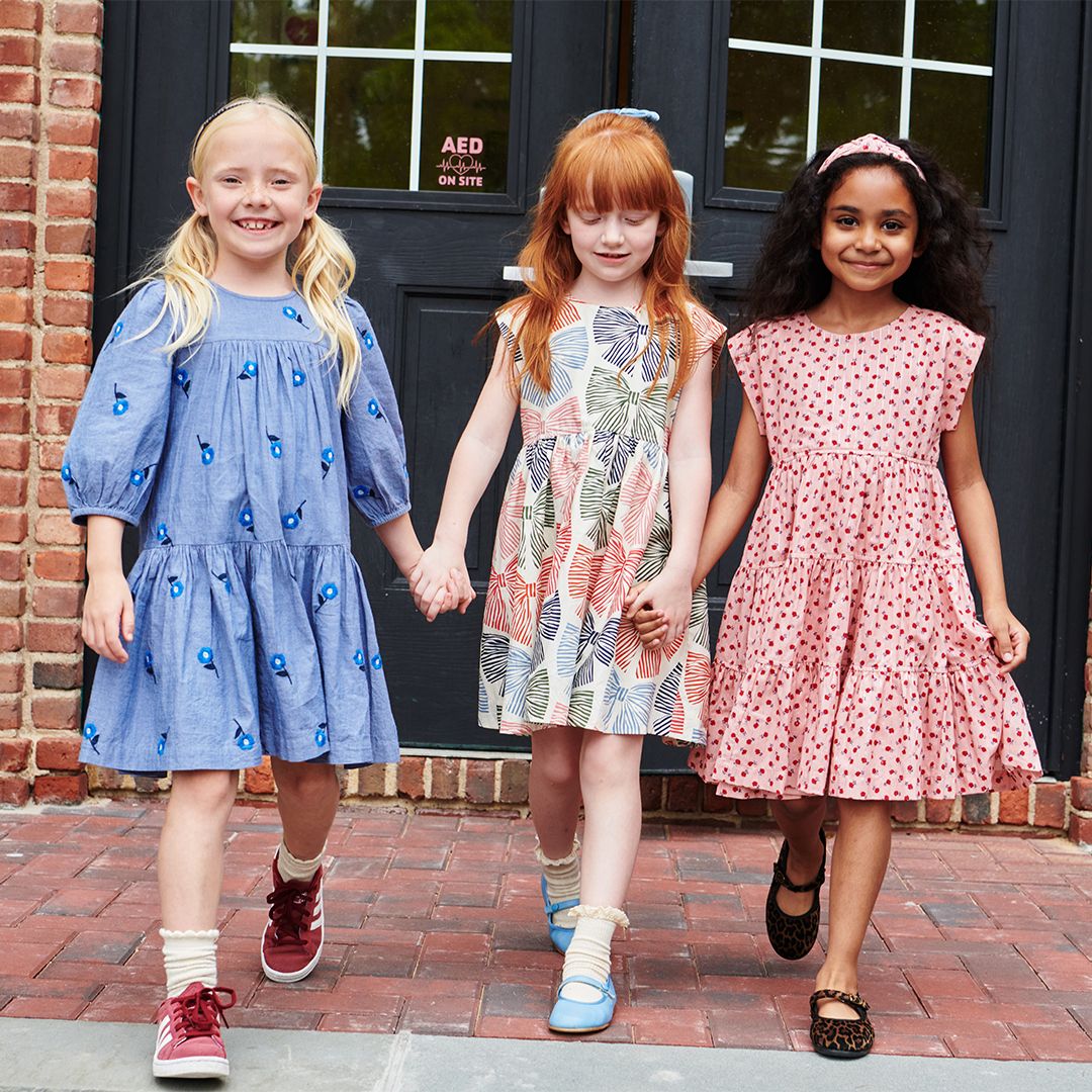 Three young girls smile at the camera as they walk hand in hand on a brick sidewalk; one wears a Pink Chicken dress with tiny pink roses, and another wears the Pink Chicken Girls' Peachy Dress.