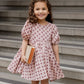 A young girl in a Rylee & Cru Baby Girls' and Girls' Rose Dress stands on stone steps, smiling and holding a closed book with both hands.