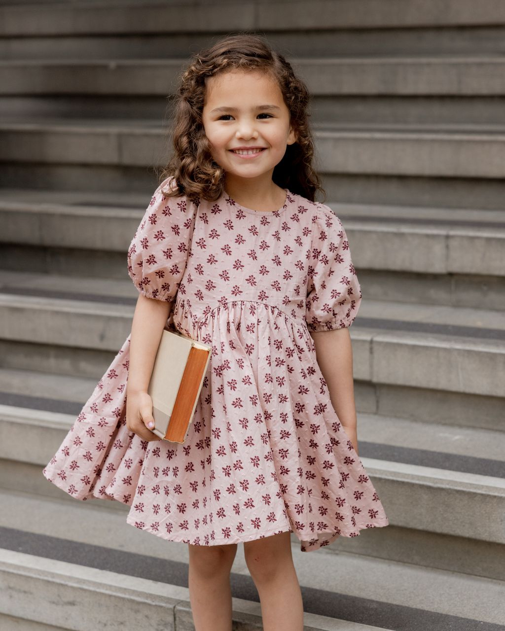 A young girl in a Rylee & Cru Baby Girls' and Girls' Rose Dress stands on stone steps, smiling and holding a closed book with both hands.