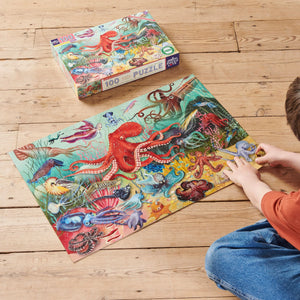 A child assembles the Eeboo Love of Octopuses & Friends 100 Piece Puzzle, featuring vibrant ocean creatures, with the puzzle box beside them on a wooden floor.