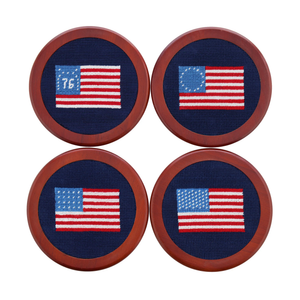 The Smathers & Branson American Flag Coasters set features four round, hand-stitched needlepoint coasters showcasing embroidered American flags—two with historic flag designs and two with the modern stars and stripes—all set in wooden frames. The set also includes a wooden coaster holder for stylish storage.