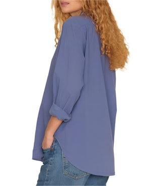 Person with curly hair wearing a loose-fitting, long-sleeve Xirena Beau Shirt by Xirena and blue jeans, standing with one hand in their pocket, facing away from the camera.