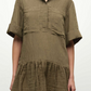 A person wears the Simkhai Cris Short Sleeve Shirt Dress in olive green, featuring a gathered skirt and textured fabric, standing against a plain background.