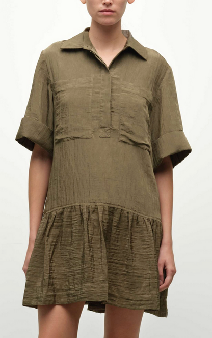 A person wears the Simkhai Cris Short Sleeve Shirt Dress in olive green, featuring a gathered skirt and textured fabric, standing against a plain background.