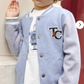 A young boy wearing the Tartine et Chocolat Baby Boys’ and Boys’ Easy Bomber Cardigan in light blue and gray with "T&C" stands facing the camera, one hand raised, showing off a sporty modern style.