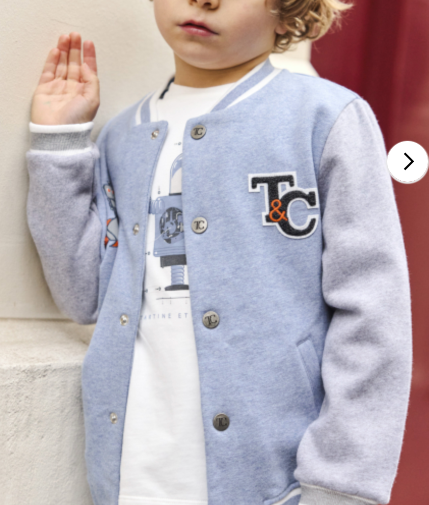 A young boy wearing the Tartine et Chocolat Baby Boys’ and Boys’ Easy Bomber Cardigan in light blue and gray with "T&C" stands facing the camera, one hand raised, showing off a sporty modern style.