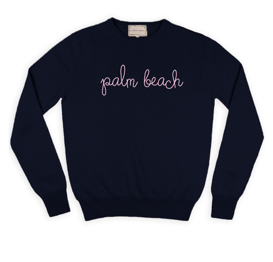 The Palm Beach Classic Longsleeve Crewneck by Lingua Franca is a navy blue cashmere sweater, hand-embroidered with "palm beach" in pink cursive across the chest, crafted from sustainably sourced cashmere.