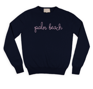 The Palm Beach Classic Longsleeve Crewneck by Lingua Franca is a navy blue cashmere sweater, hand-embroidered with "palm beach" in pink cursive across the chest, crafted from sustainably sourced cashmere.