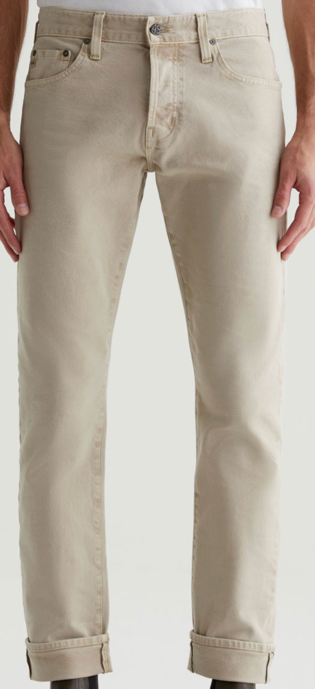 A person wearing light beige Tellis Selvage jeans by AG Jeans, featuring rolled cuffs and crafted from Japanese stretch selvage denim, shown waist to ankle against a plain background.