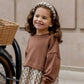 A young girl with curly hair smiles outdoors by a bicycle basket, wearing a Rylee & Cru padded headband, brown sweatshirt, and floral skirt.