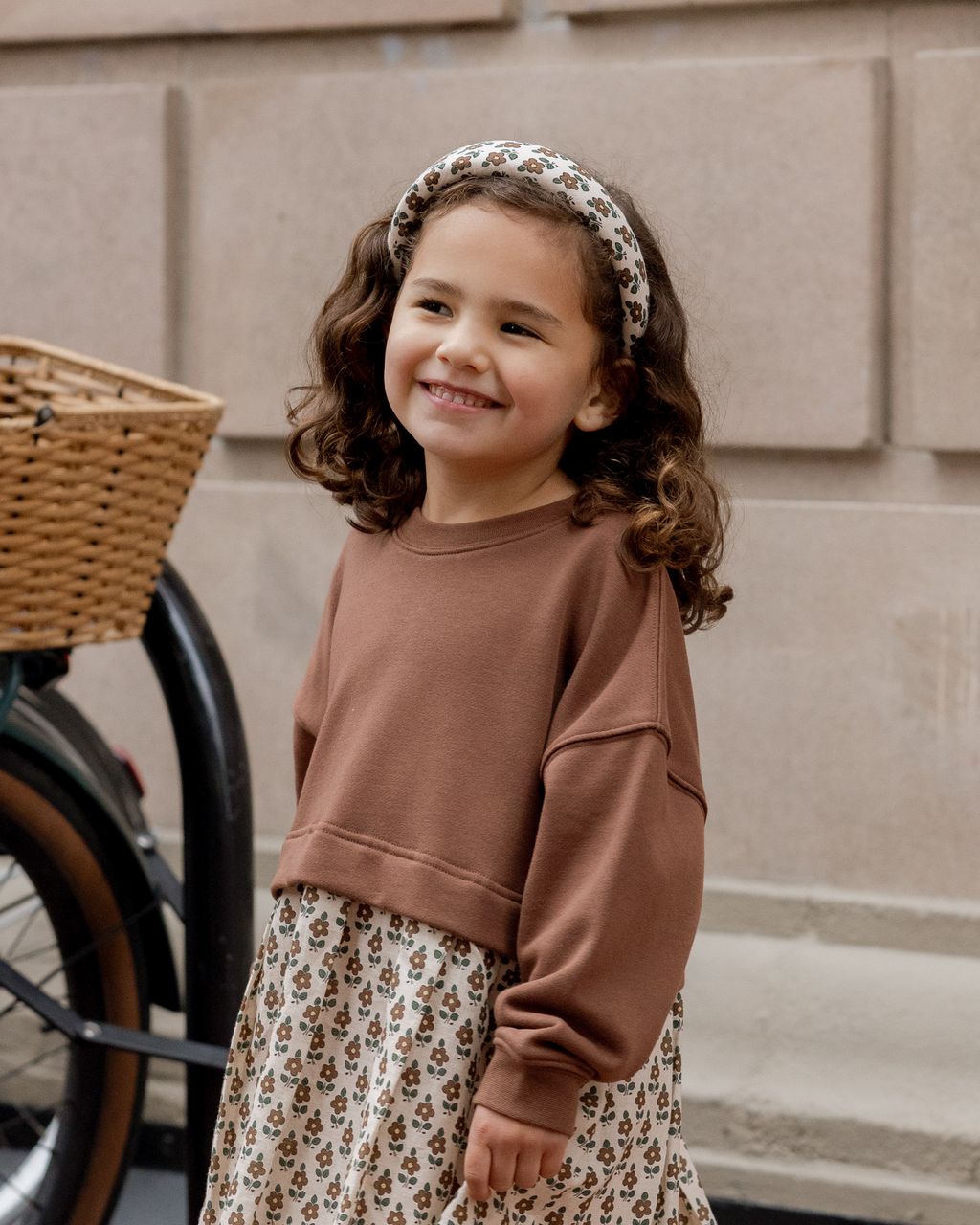A young girl with curly hair smiles outdoors by a bicycle basket, wearing a Rylee & Cru padded headband, brown sweatshirt, and floral skirt.